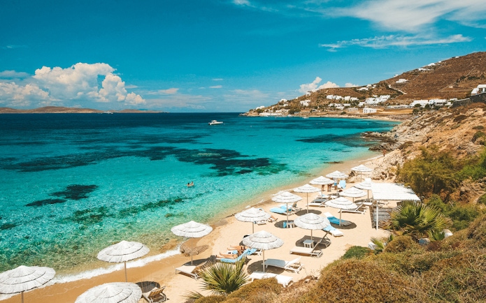 Mykonos beach with umbrellas and loungers, clear blue sea, and distant hills.