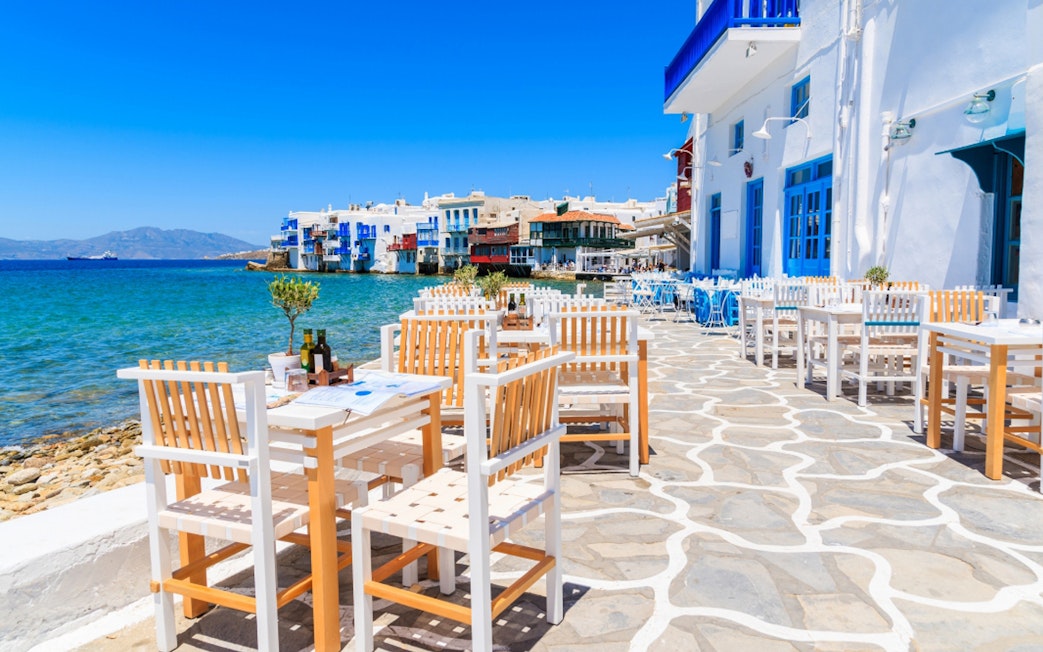 Outdoor dining area by the sea in Mykonos, Greece, with white and blue buildings.