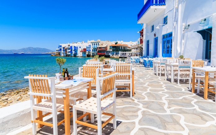 Outdoor dining area by the sea in Mykonos, Greece, with white and blue buildings.