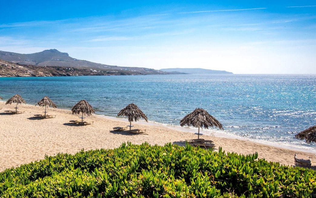 Mykonos beach with straw umbrellas and clear blue sea on a sunny day.
