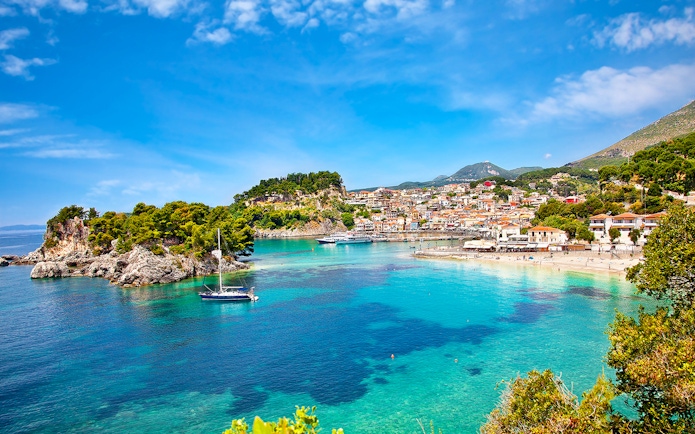 Sailboat in turquoise waters near Parga, Greece, with coastal town and lush hills in the background.