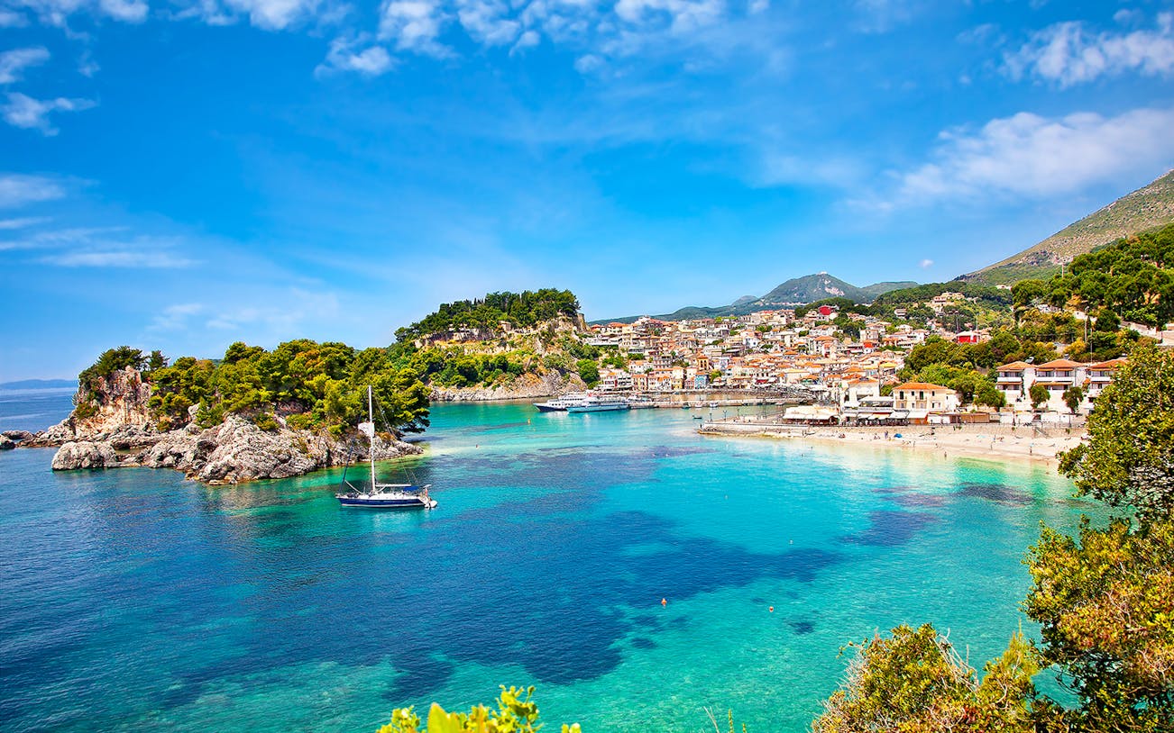Sailboat in turquoise waters near Parga, Greece, with coastal town and lush hills in the background.