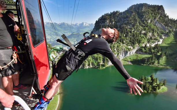 Person bungy jumping from gondola at Stockhorn, Switzerland, over a scenic lake and mountain landscape.