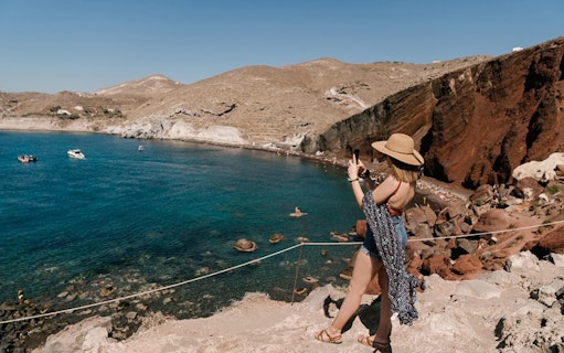 Tourist taking photo of Santorini's Red Beach cliffs and blue sea during volcano sightseeing tour.