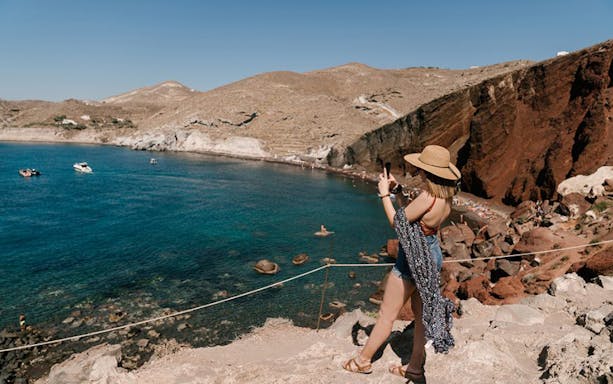 Tourist taking photo of Santorini's Red Beach cliffs and blue sea during volcano sightseeing tour.