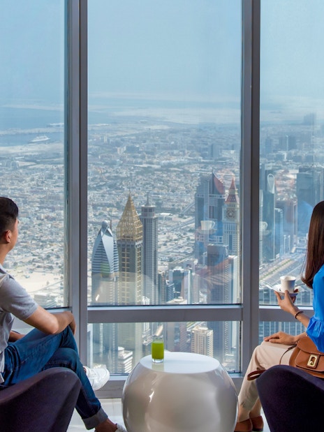 Visitors enjoying city view from Burj Khalifa café with coffee, Dubai skyline visible.