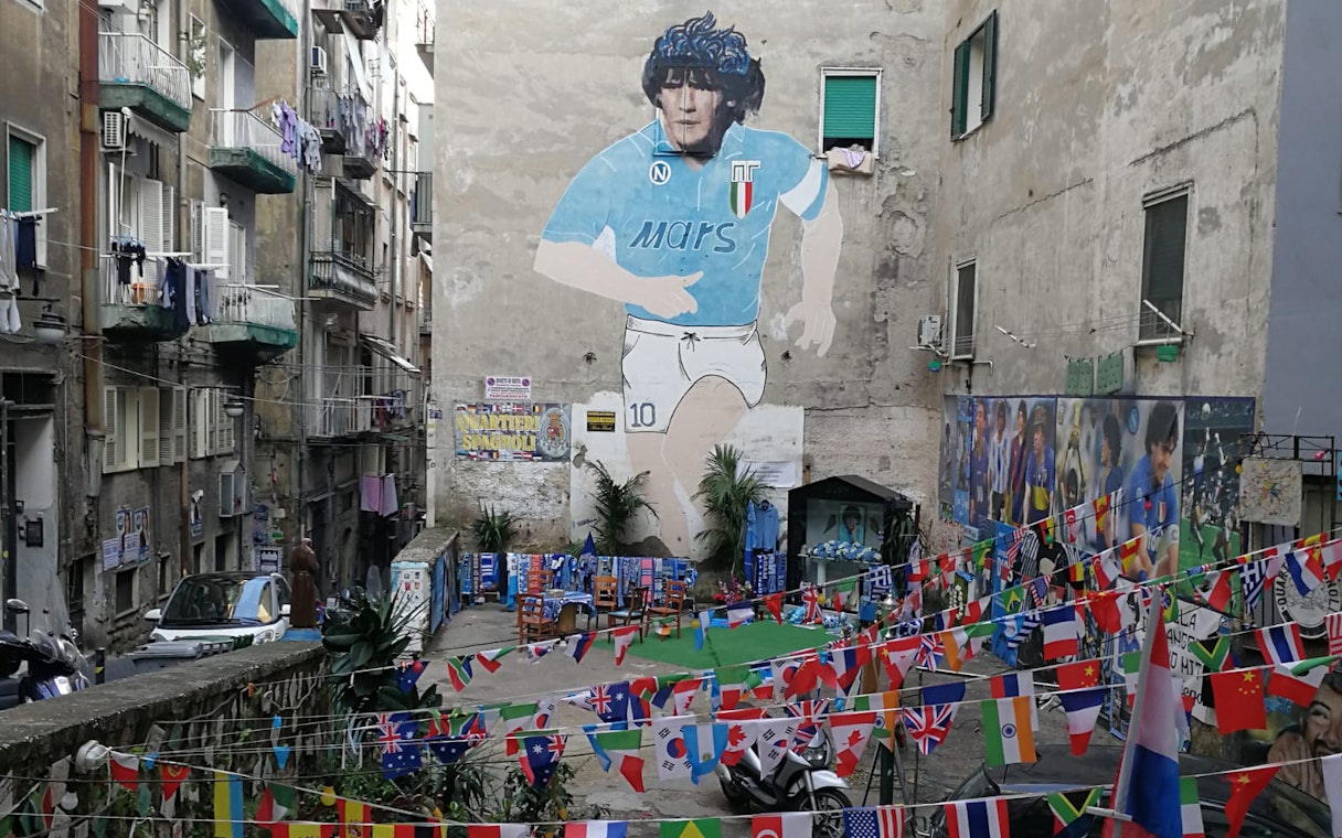 Mural of Diego Maradona in a Naples alley with international flags and memorabilia.