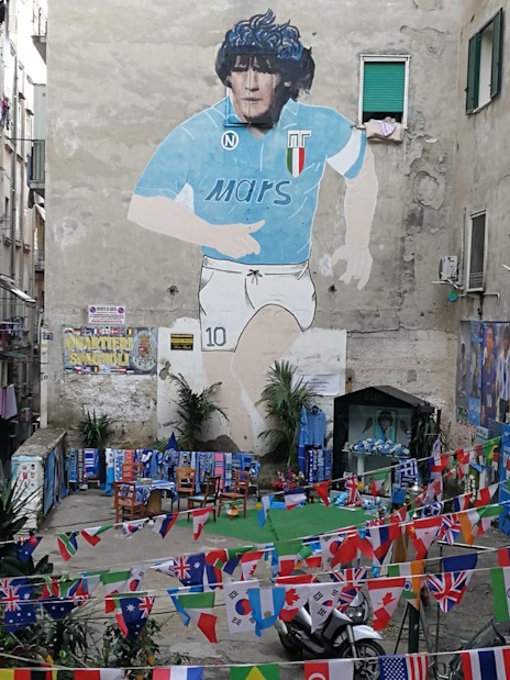Mural of Diego Maradona in a Naples alley with international flags and memorabilia.