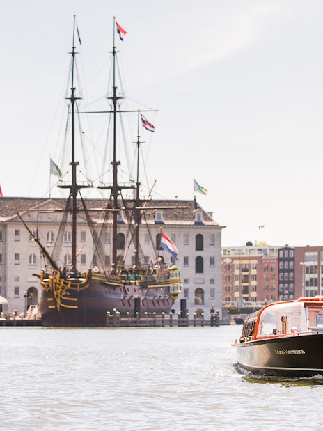 Amsterdam canal cruise boat near NEMO Science Museum with historic ship in background.