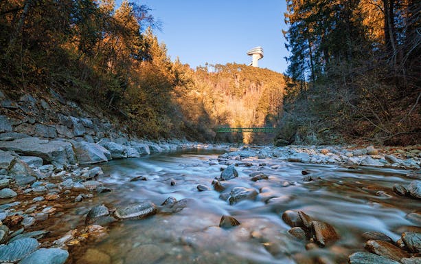 Bergisel Stadium tower above a forested hill with a rocky stream in the foreground.