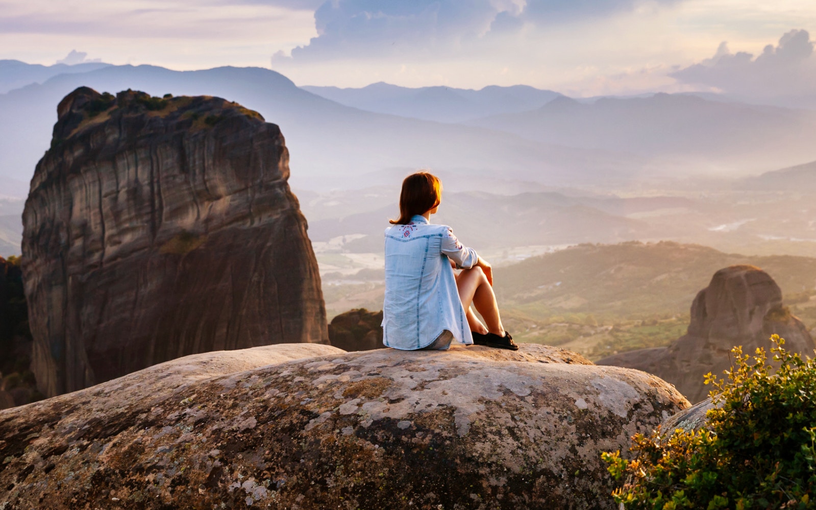 Person sitting on rock overlooking Meteora rock formations at sunset, Greece.