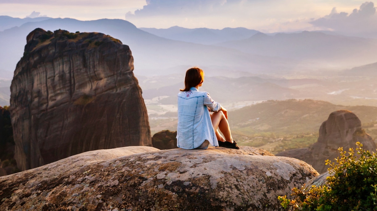 Person sitting on rock overlooking Meteora rock formations at sunset, Greece.