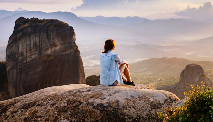 Person sitting on rock overlooking Meteora rock formations at sunset, Greece.