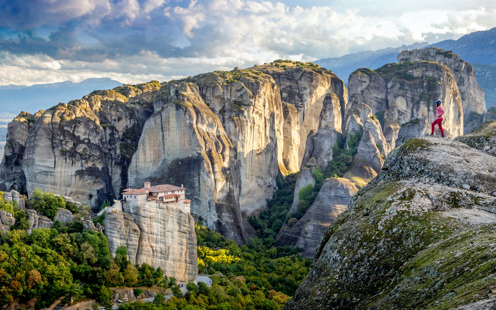 Meteora rock formations with a monastery and a person standing on a cliff, Greece.