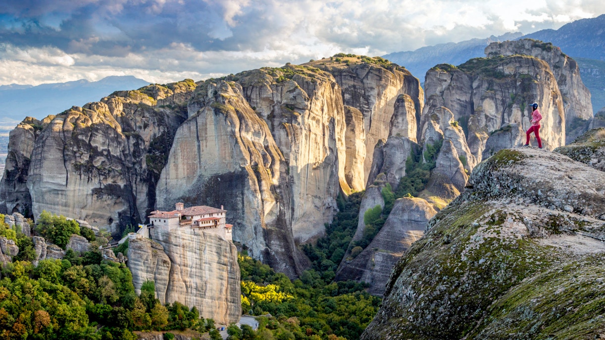 Meteora rock formations with a monastery and a person standing on a cliff, Greece.