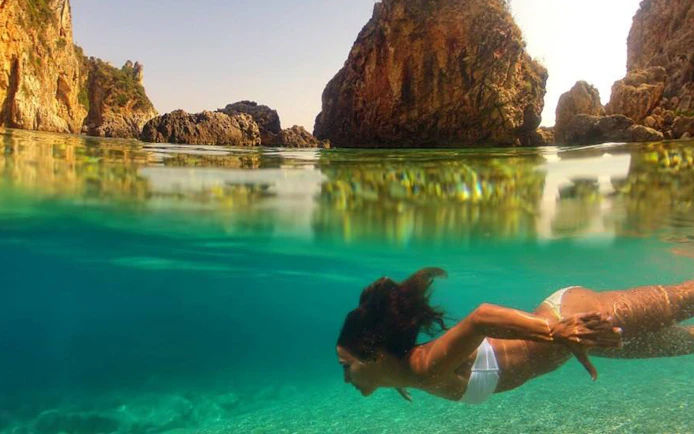 Snorkeler exploring clear waters near rocky cliffs in Corfu during a full-day boat trip with BBQ.