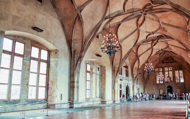 Prague Castle interior with vaulted ceilings and chandeliers, tourists exploring the hall.