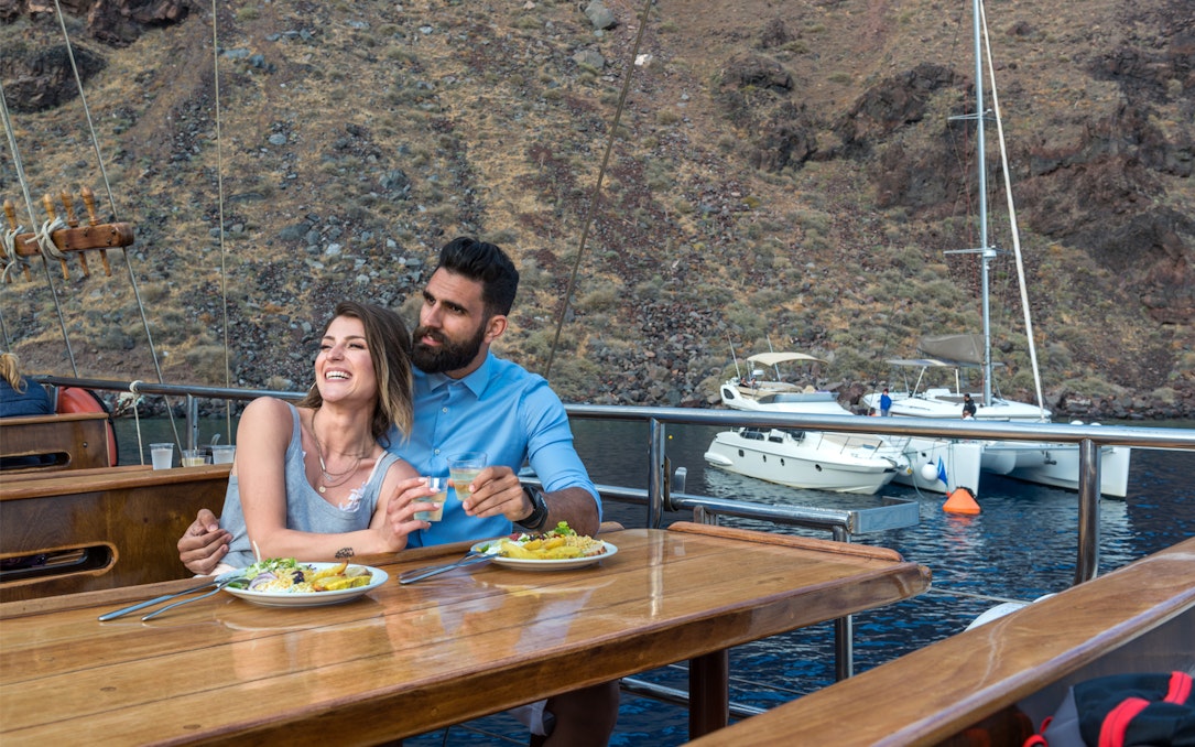 Couple enjoying dinner on a Santorini sunset cruise with a view of volcanic islands.