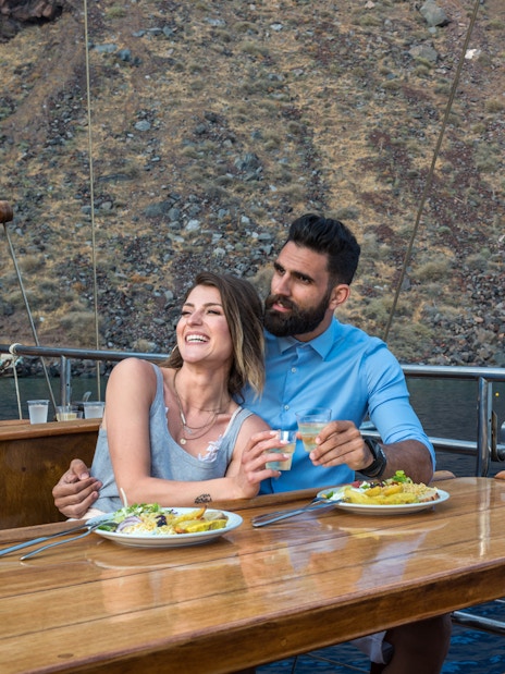 Couple enjoying dinner on a Santorini sunset cruise with a view of volcanic islands.