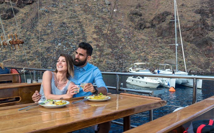 Couple enjoying dinner on a Santorini sunset cruise with a view of volcanic islands.