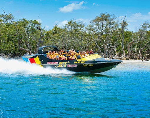 Jet boat with passengers speeding on water near a forested shoreline.