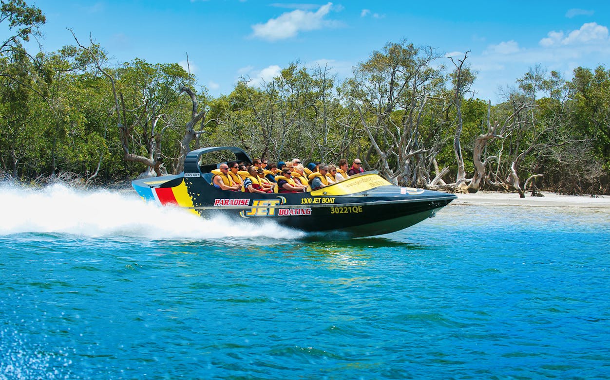 Jet boat speeding on water with passengers wearing life jackets, surrounded by trees.