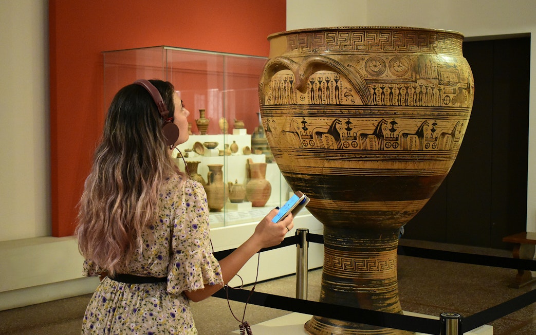 Visitor using audio guide at National Archaeological Museum, viewing ancient Greek pottery.