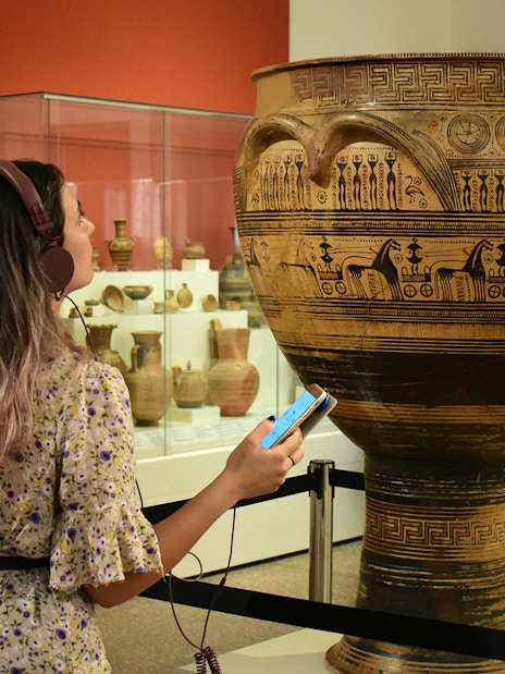 Visitor using audio guide at National Archaeological Museum, viewing ancient Greek pottery.