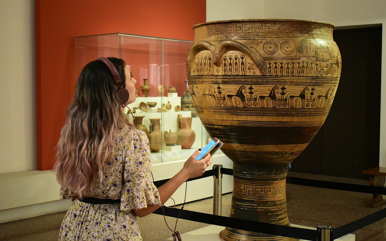 Visitor using audio guide at National Archaeological Museum, viewing ancient Greek pottery.