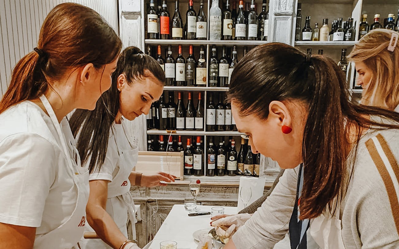 Women preparing pasta dishes in a cooking class with wine bottles in the background.