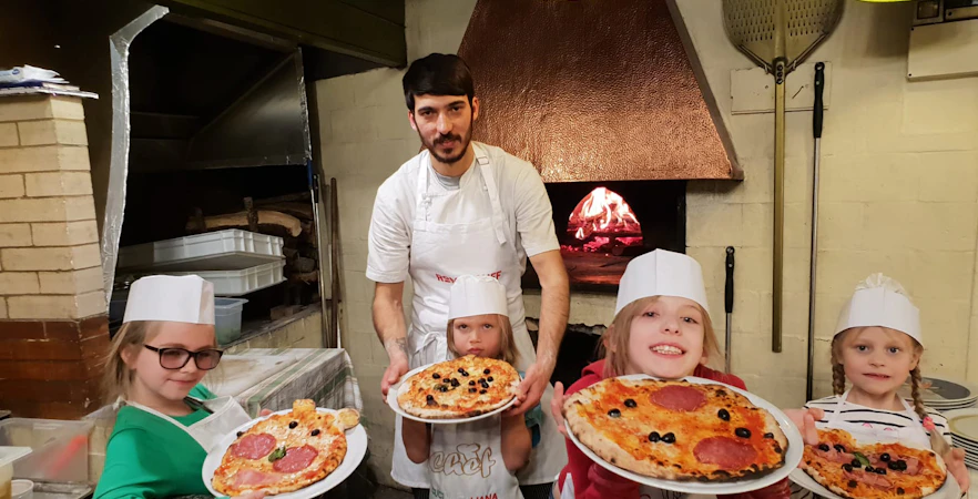 Children and instructor holding pizzas in a wood-fired oven class.