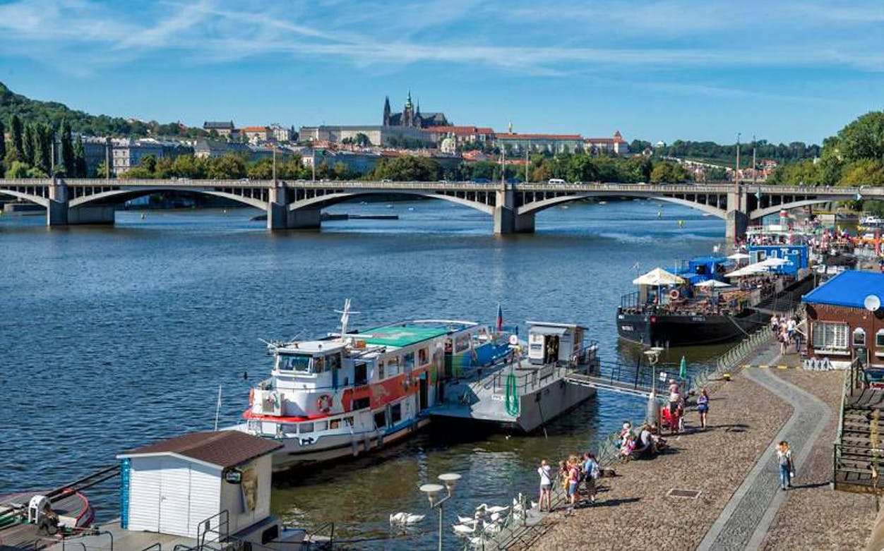Cruise boat docked by a river with a bridge and cityscape in the background, Prague.