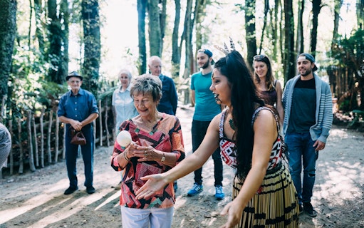 Elderly woman learning poi dance at Tamaki Maori Village, surrounded by group in forest setting.