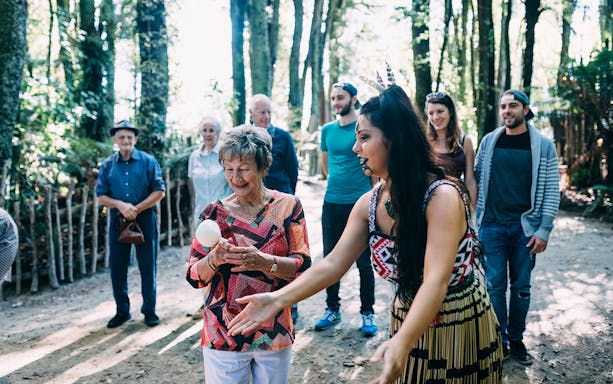Elderly woman learning poi dance at Tamaki Maori Village, surrounded by group in forest setting.