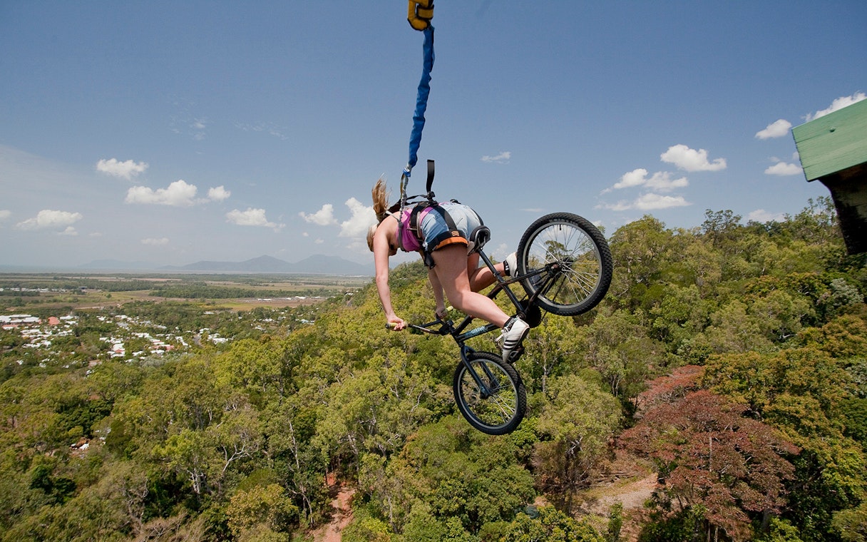 Person bungy jumping with a bicycle over lush landscape in Cairns, Australia.