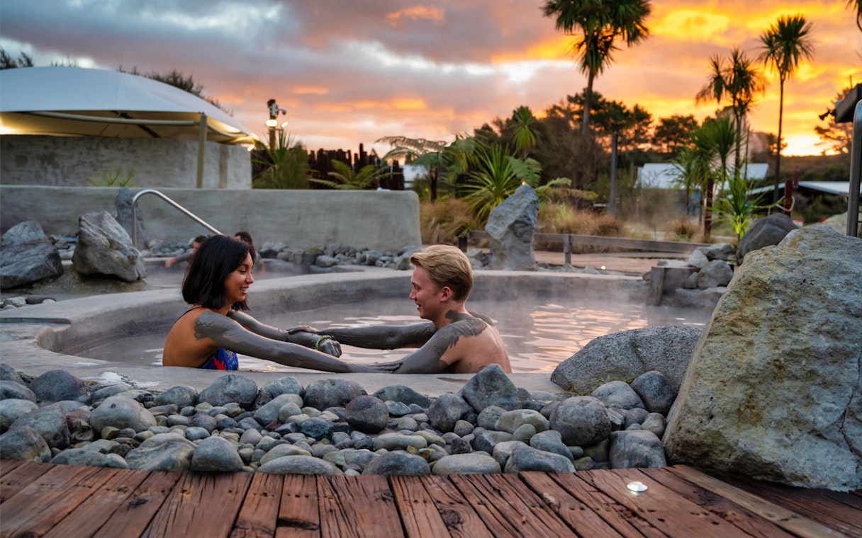 Couple enjoying mud bath at Hell's Gate Spa, New Zealand, with sunset backdrop.