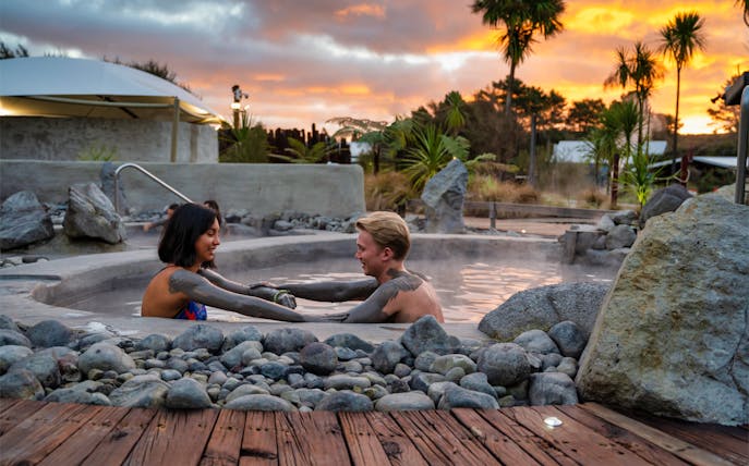 Couple enjoying mud bath at Hell's Gate Spa, New Zealand, with sunset backdrop.