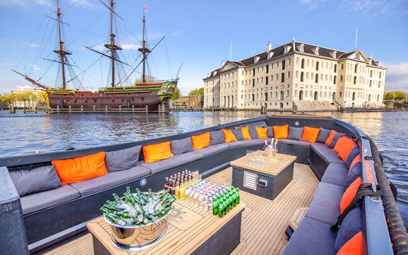 Luxury open boat with drinks cruising Amsterdam canals near historic ship and museum.
