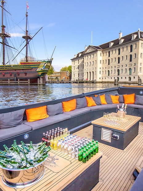 Luxury open boat with drinks cruising Amsterdam canals near historic ship and museum.