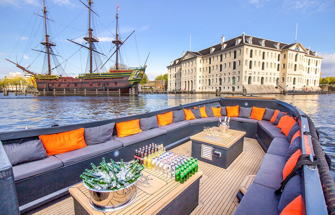 Luxury open boat with drinks cruising Amsterdam canals near historic ship and museum.