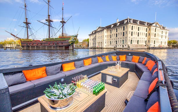 Luxury open boat with drinks cruising Amsterdam canals near historic ship and museum.
