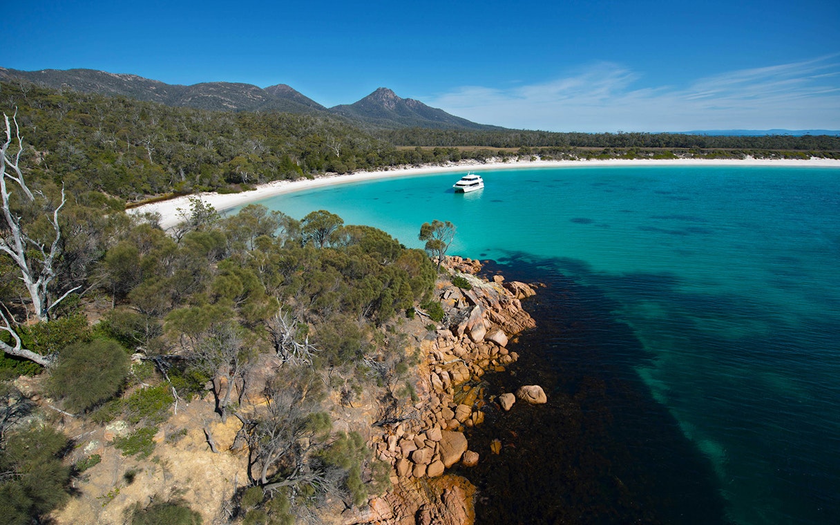 Cruise boat in turquoise waters of Wineglass Bay, Tasmania, with forested hills and rocky shoreline.