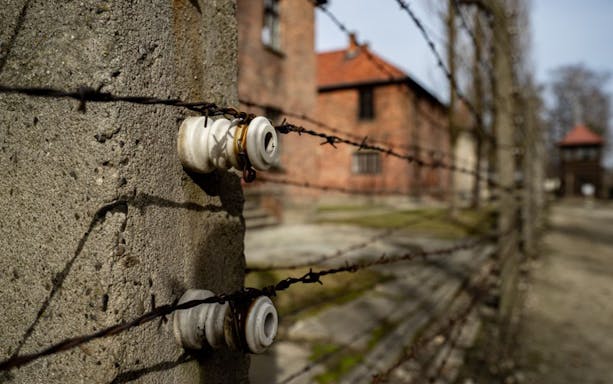 Barbed wire fence at Auschwitz-Birkenau with brick buildings in the background.