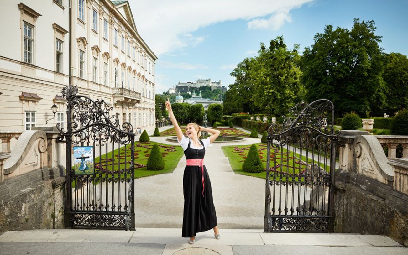 Woman in traditional dress at Mirabell Gardens, Salzburg, with Hohensalzburg Fortress in the background.
