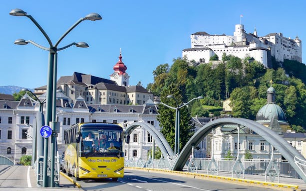 Hop-on hop-off bus crossing a bridge in Salzburg with Hohensalzburg Fortress in the background.