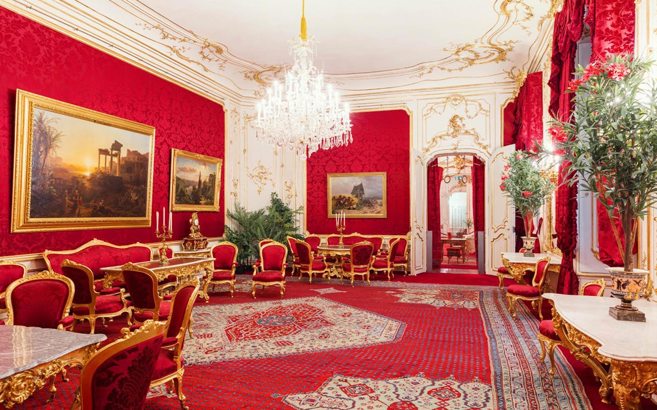 Ornate room with red and gold decor in Vienna palace, featuring chandeliers and classic paintings.