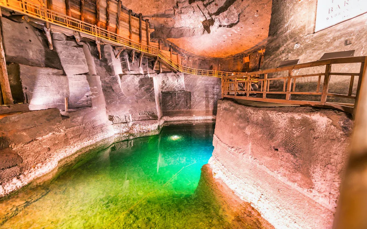 Underground salt mine chamber with illuminated green water pool and wooden walkways.