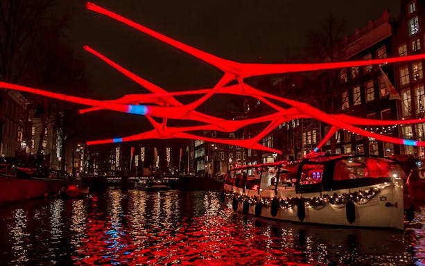 Luxury canal cruise under illuminated red art installation at Amsterdam Light Festival.