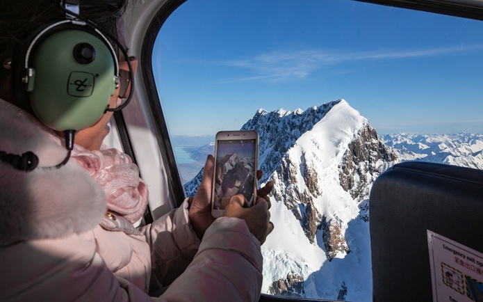 Passenger photographing snow-covered peaks during scenic flight over Fox and Franz Josef Glaciers.