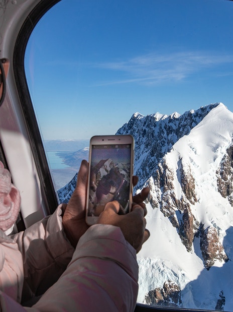 Passenger photographing snow-covered peaks during scenic flight over Fox and Franz Josef Glaciers.